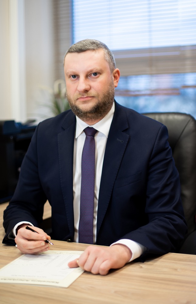 Mężczyzna w garniturze siedzi przy biurku, trzyma długopis nad dokumentem z tabelą, patrząc wprost w obiektyw.