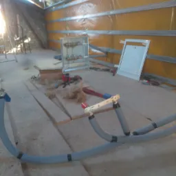 Poddasze w trakcie prac adaptacyjnych: widoczne rury instalacji grzewczej z czarnymi opaskami, narzędzia, płyty OSB oraz żółta folia izolacyjna przymocowana do metalowych profili.