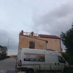 Renowacja dachu budynku z ceglanymi kominami, widoczna częściowo nowa dachówka i drewniana konstrukcja, na pierwszym planie samochód dostawczy z reklamą usług ciesielsko-dekarskich.