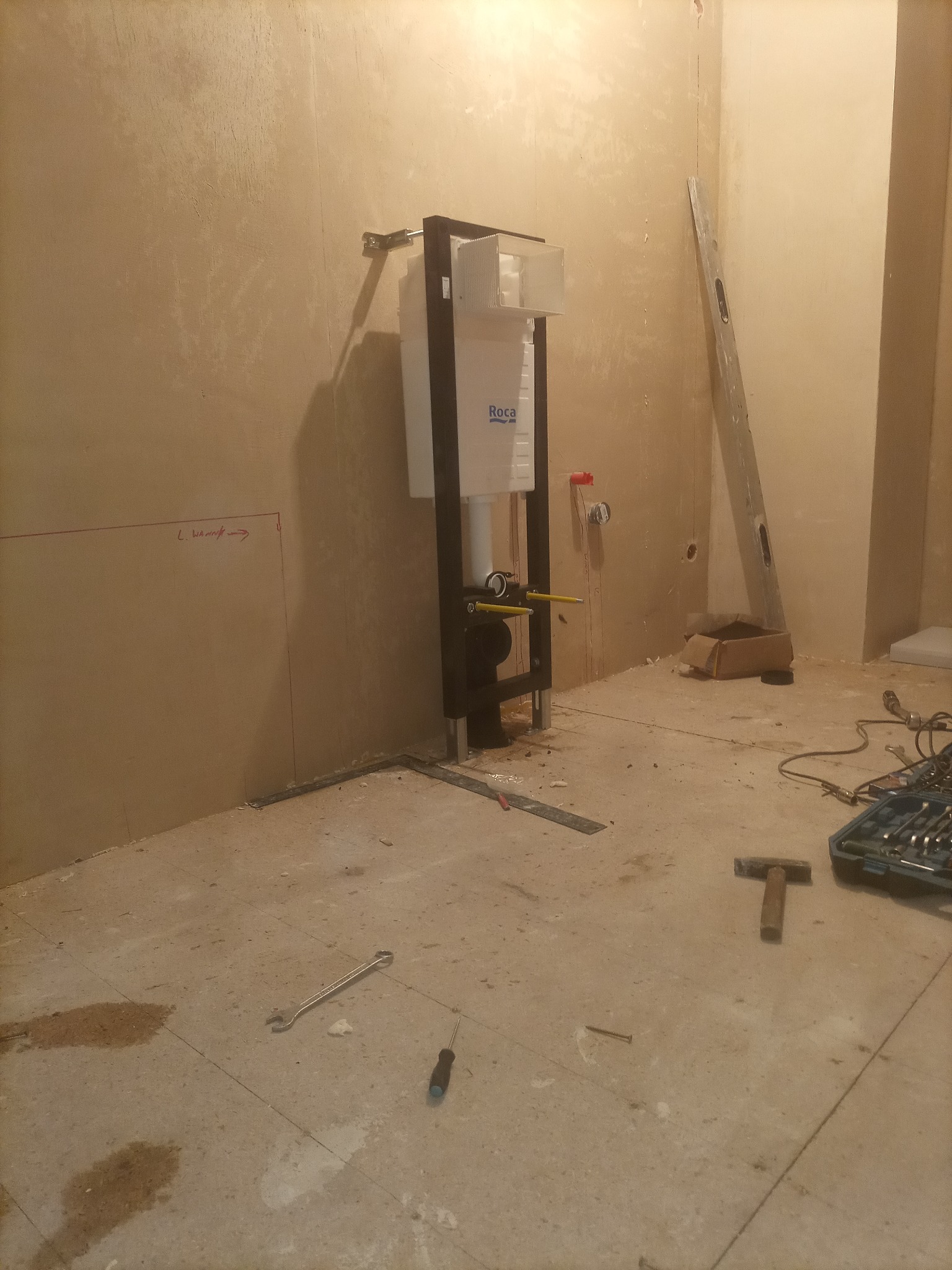 Montaż stelaża podtynkowego WC marki Roca w surowym wnętrzu łazienki, widoczne narzędzia i poziomica, ściany w trakcie przygotowania do wykończenia.