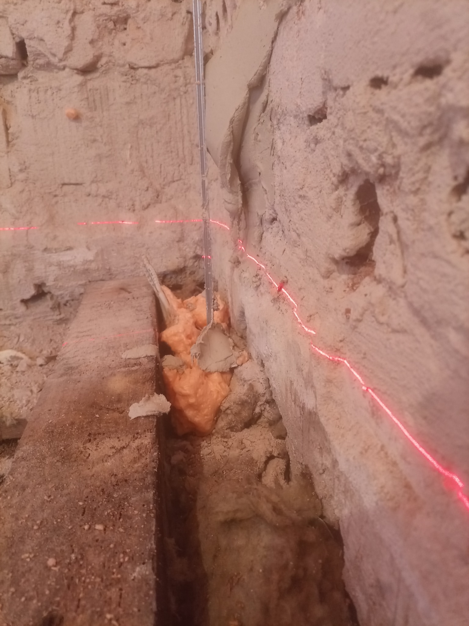 Fragment ściany z nierówną powierzchnią, wypełniony pianką montażową i zaprawą, wyznaczony czerwoną linią lasera.