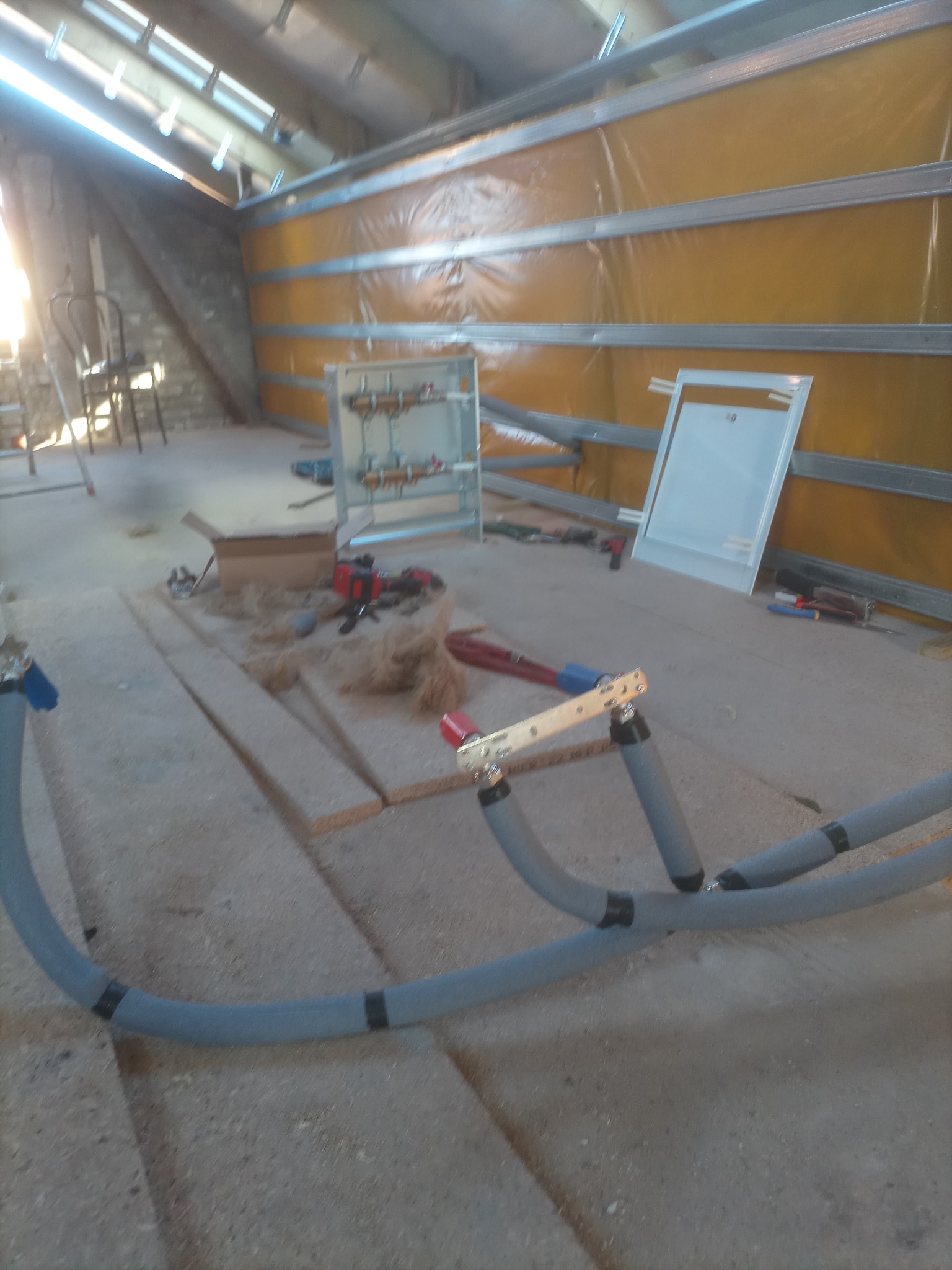 Poddasze w trakcie prac adaptacyjnych: widoczne rury instalacji grzewczej z czarnymi opaskami, narzędzia, płyty OSB oraz żółta folia izolacyjna przymocowana do metalowych profili.