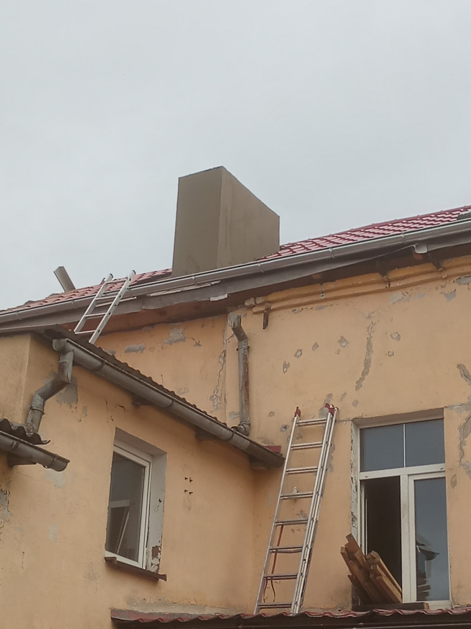 Dwupiętrowy budynek z żółtą elewacją w trakcie prac remontowych, z drabiny opartymi o dach i ścianę, widoczny komin z nową obudową, oraz fragment dachu z rynną i nową dachówką.