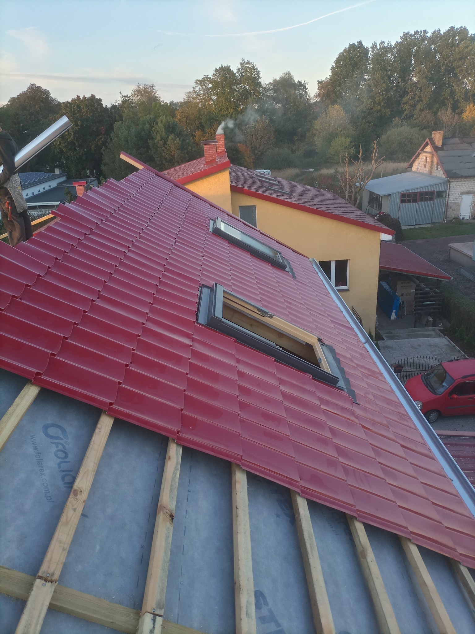 Montaż czerwonej blachodachówki z oknami dachowymi na konstrukcji drewnianej z widoczną membraną dachową, w tle zabudowa jednorodzinna i drzewa.