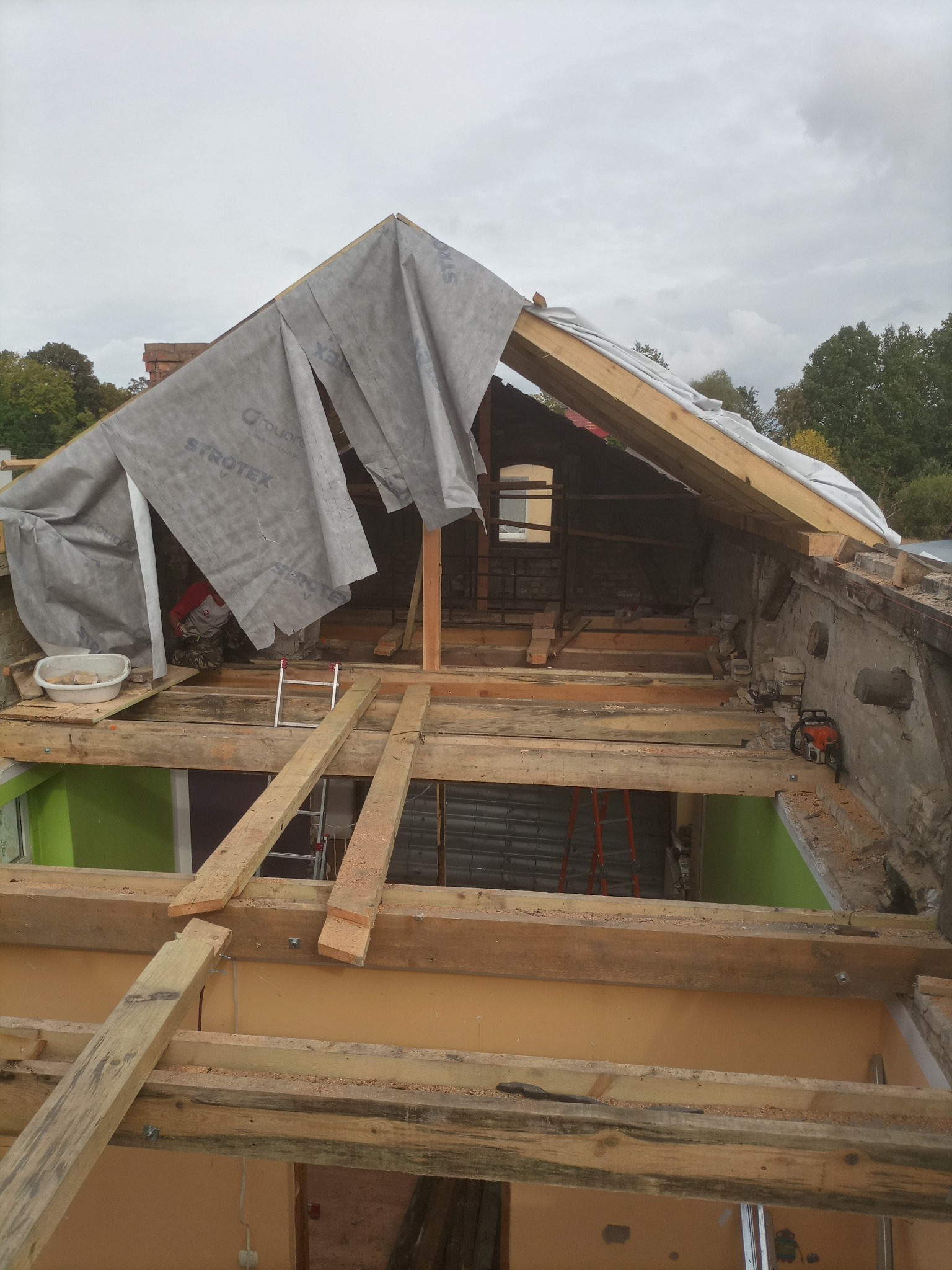 Konstrukcja dachu w trakcie remontu z widoczną więźbą dachową, folią ochronną Strotex i fragmentem muru z piłą łańcuchową.