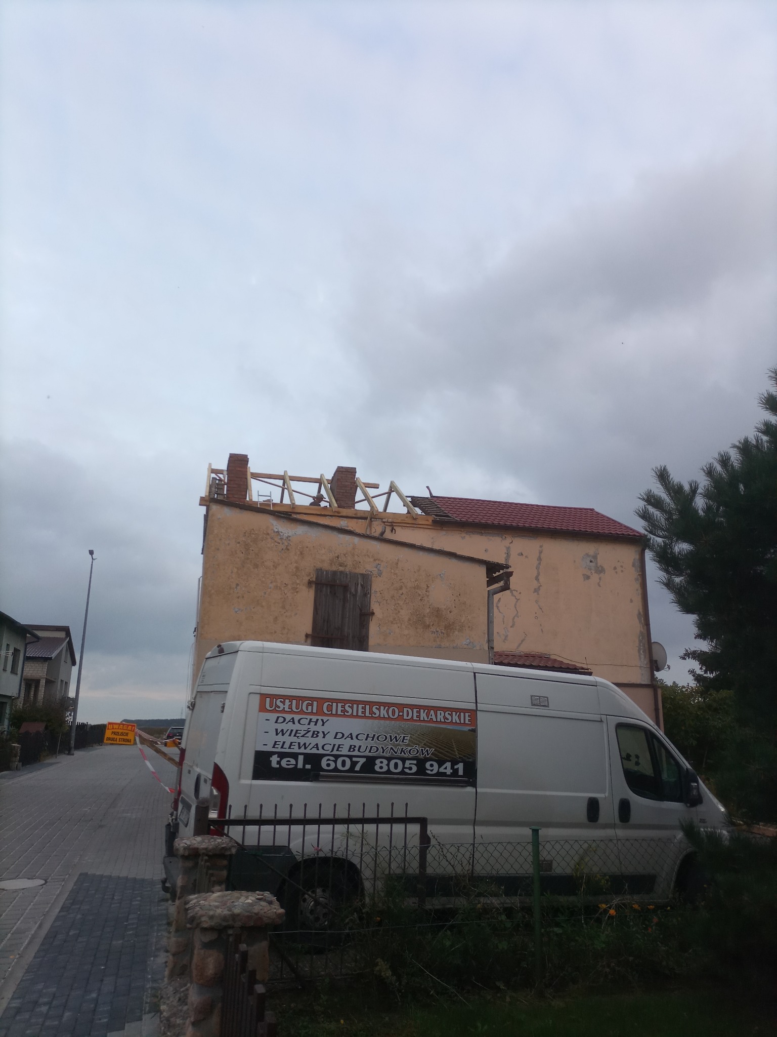 Renowacja dachu budynku z ceglanymi kominami, widoczna częściowo nowa dachówka i drewniana konstrukcja, na pierwszym planie samochód dostawczy z reklamą usług ciesielsko-dekarskich.