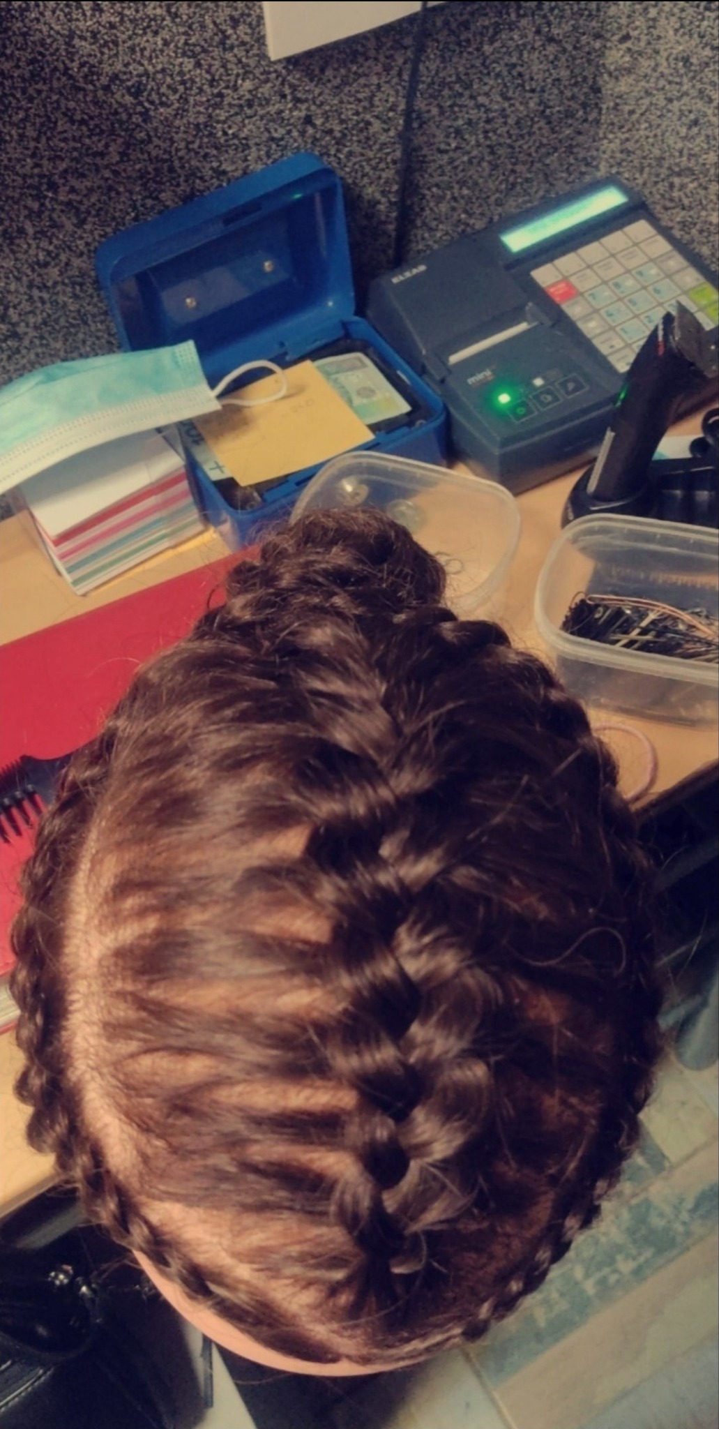 Góra głowy z ciemnymi, splecionymi włosami na tle biurka z kasą fiskalną, niebieskim pudełkiem i akcesoriami.