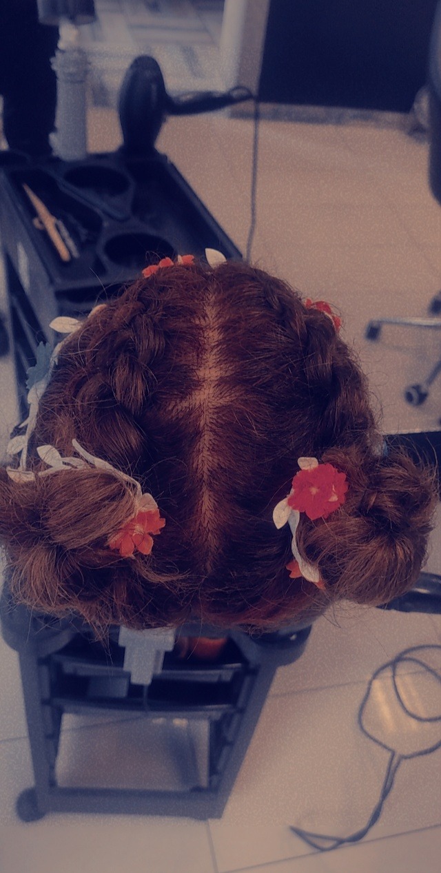 Fryzura z warkoczami i czerwonymi kwiatami, widok z góry na głowę manekina fryzjerskiego, tło rozmyte z widocznym wózkiem fryzjerskim z narzędziami.