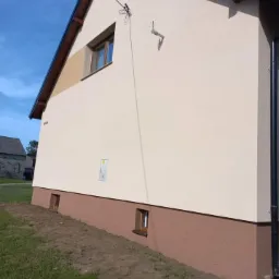 Elewacja domu jednorodzinnego po remoncie, z widocznym ociepleniem i nowym kolorem, z brązowym cokołem i widocznymi małymi okienkami piwnicznymi, na tle błękitnego nieba.