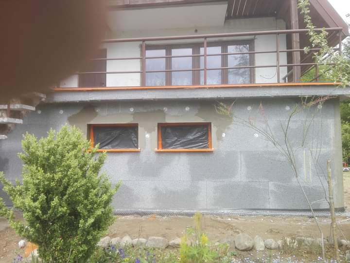 Ocieplanie ściany zewnętrznej budynku styropianem, widoczne zaślepki na styropianie, okna zabezpieczone folią, na dole rabata kwiatowa.