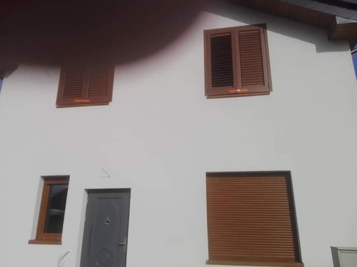 Biała fasada domu z brązowymi okiennicami, oknem i drzwiami wejściowymi w szarym kolorze. Widoczne lekkie pęknięcia na ścianie.