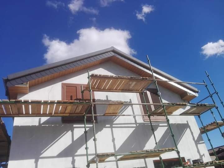 Biały dom jednorodzinny w trakcie prac remontowych, widoczne rusztowanie, brązowe okiennice, szara dachówka i błękitne niebo z chmurami.