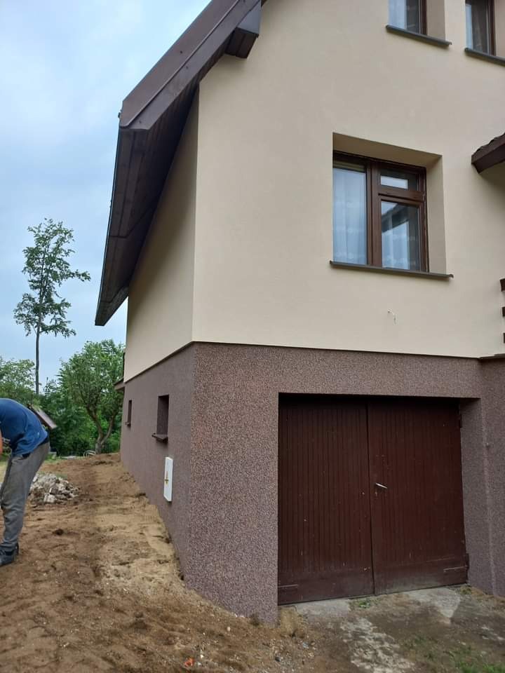 Elewacja domu jednorodzinnego z brązowym cokołem i beżową górną częścią, widoczne okna z brązowymi ramami i fragment ogrodu z pracującym mężczyzną.