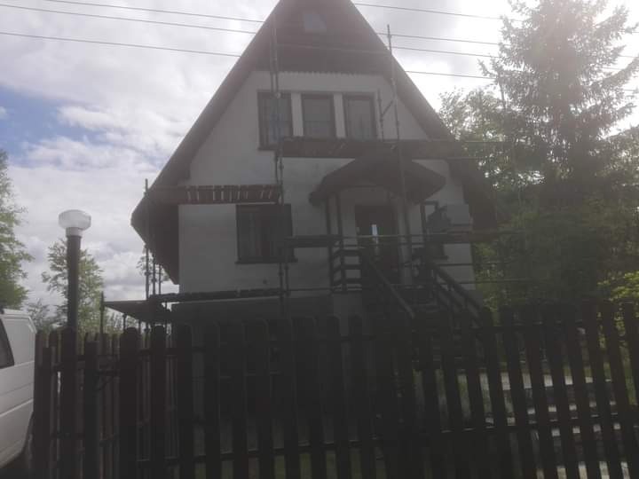 Dom jednorodzinny w trakcie prac elewacyjnych, widoczne rusztowania i świeżo nałożony tynk, częściowo zasłonięty przez drewniany płot.