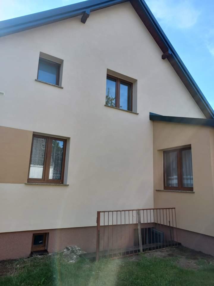 Elewacja domu jednorodzinnego po remoncie, z widocznymi oknami w drewnianych ramach i fragmentem metalowego ogrodzenia przy niewielkim oknie piwnicznym, na tle błękitnego nieba.