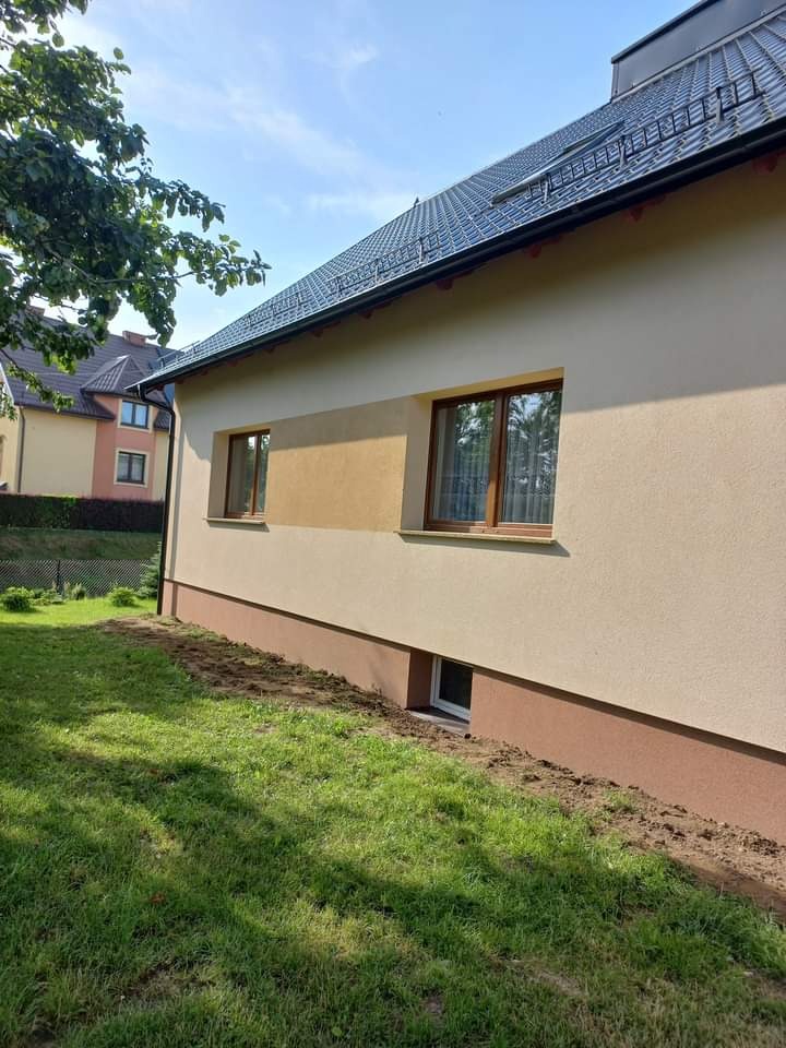 Elewacja domu jednorodzinnego w odcieniach beżu i brązu, widok z zewnątrz na świeżo wykończoną ścianę z oknami, trawnik częściowo zniszczony po pracach.