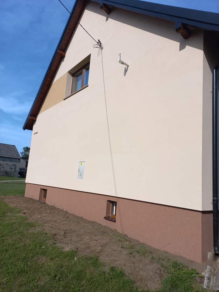 Elewacja domu jednorodzinnego po remoncie, z widocznym ociepleniem i nowym kolorem, z brązowym cokołem i widocznymi małymi okienkami piwnicznymi, na tle błękitnego nieba.