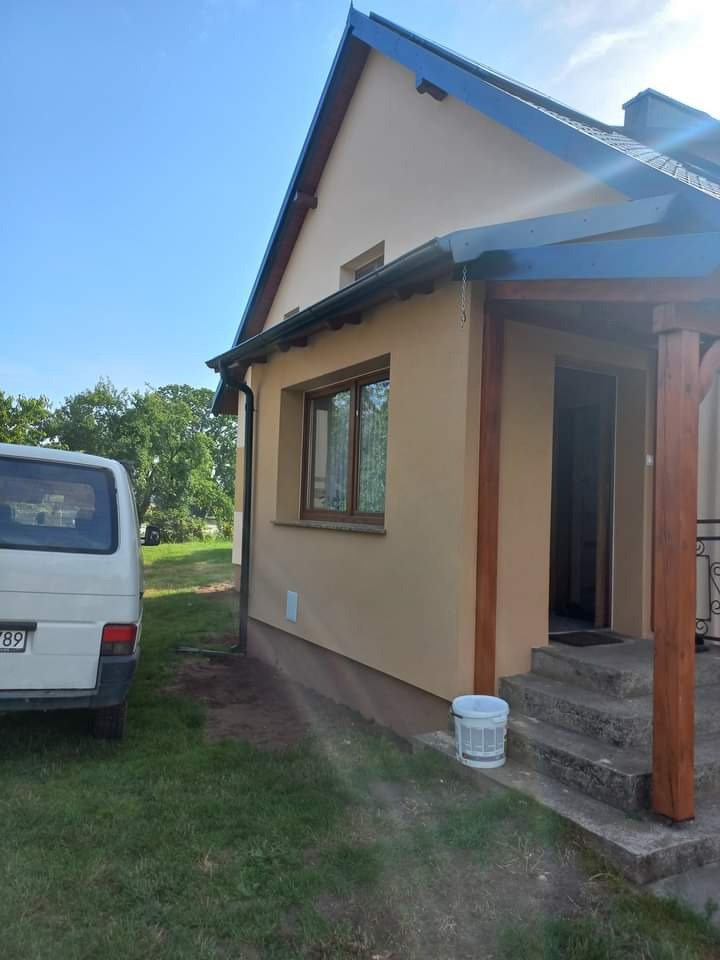 Elewacja domu jednorodzinnego w Kartuzach po nałożeniu tynku, widoczny fragment samochodu dostawczego i wiadro na schodach.