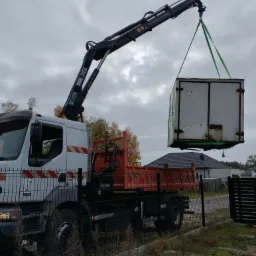 Żuraw samochodowy podnosi kontener na tle pochmurnego nieba i domów. Widoczne ogrodzenie posesji i fragment krajobrazu.