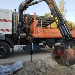 eM Lift MARCIN DĄBROWSKI - Pomarańczowa wywrotka z dźwigiem kopie ziemię na działce. Widoczne podpory stabilizujące pojazd. Ziemia w wykopie i na pryzmie. Częściowo widoczne ogrodzenie.