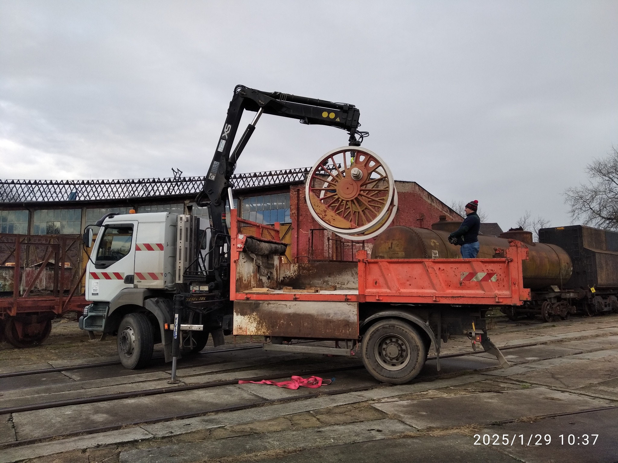 Ciężarówka z dźwigiem transportuje duże koło wagonu na tle wagonów i ceglanego budynku. Osoba w czerwonej czapce nadzoruje operację. Szyny kolejowe na ziemi.