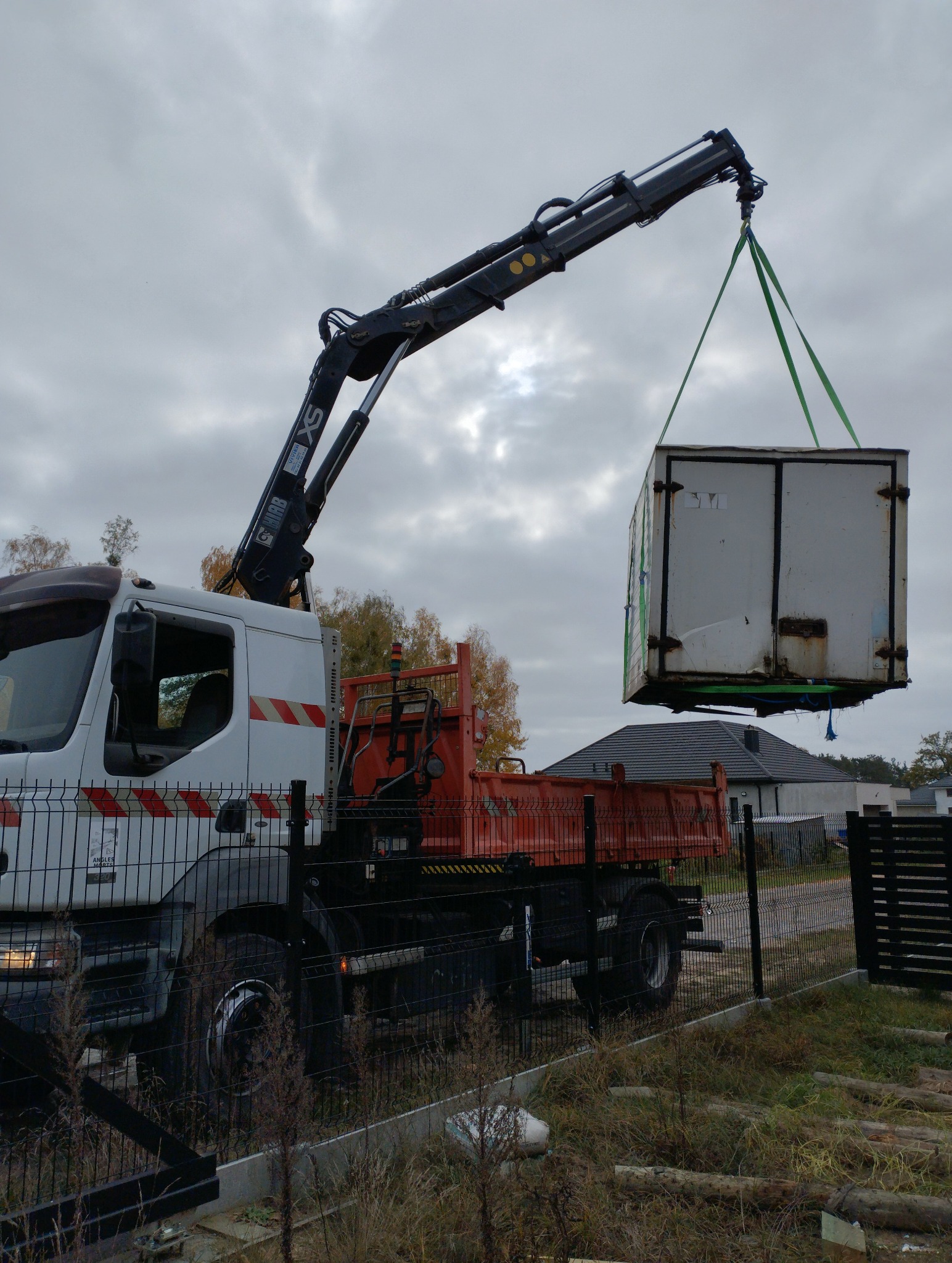 Żuraw samochodowy podnosi kontener na tle pochmurnego nieba i domów. Widoczne ogrodzenie posesji i fragment krajobrazu.