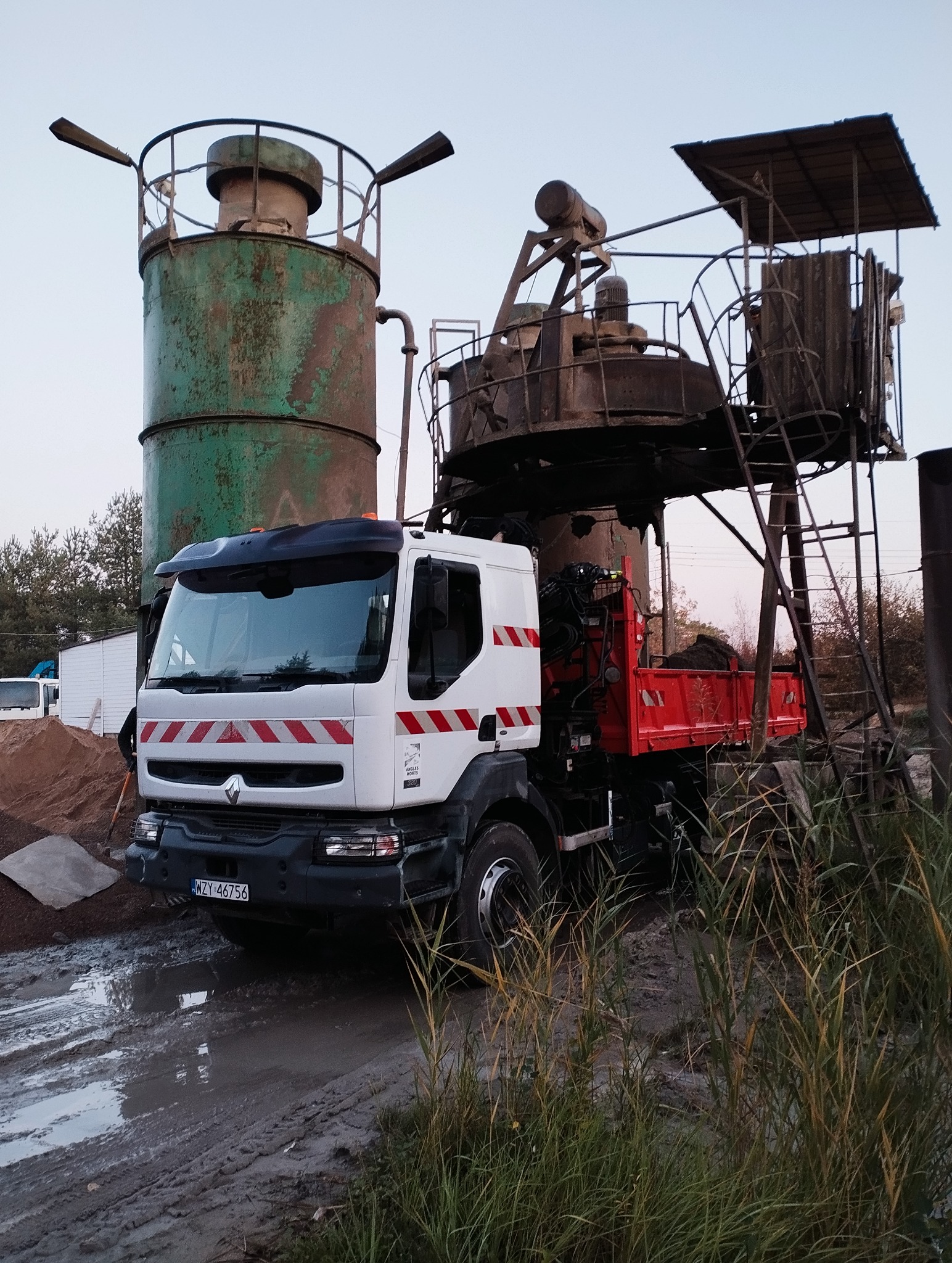 Ciężarówka Renault z czerwoną skrzynią ładunkową stoi na błotnistym terenie obok konstrukcji do mieszania materiałów budowlanych. Widoczne silosy i roślinność.