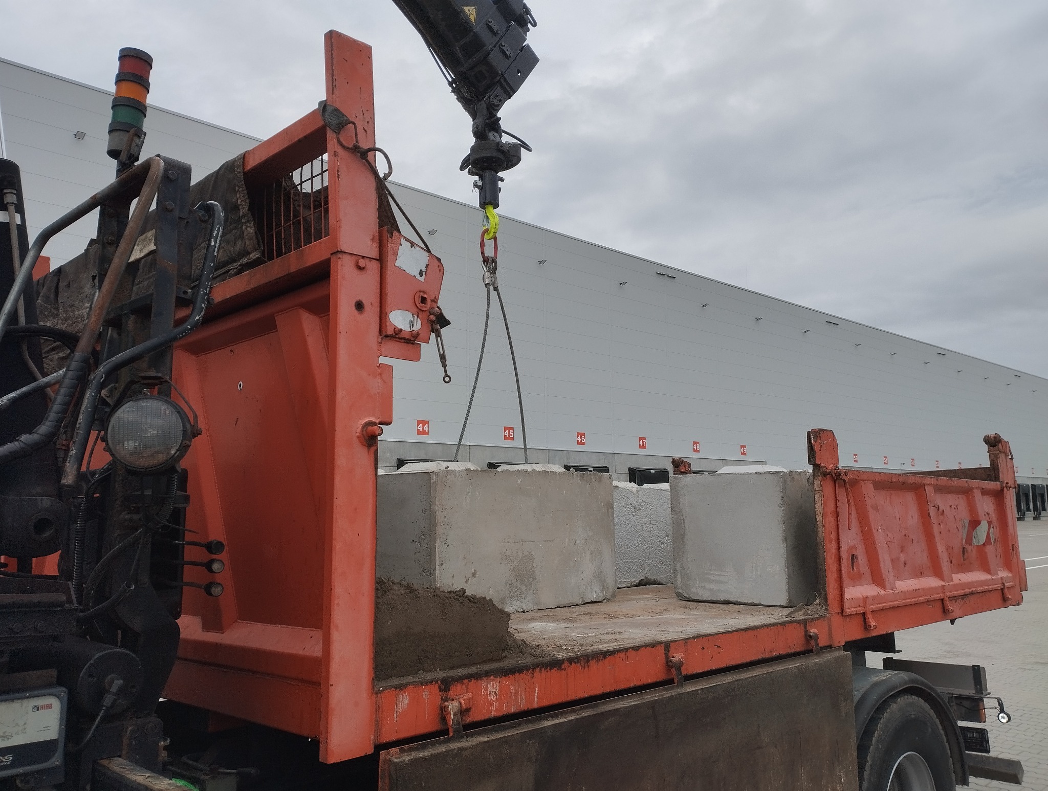 Pomarańczowa ciężarówka z dźwigiem załadowana betonowymi blokami na tle magazynu. Dźwig unosi kolejny blok. Widoczne numery ramp załadunkowych.