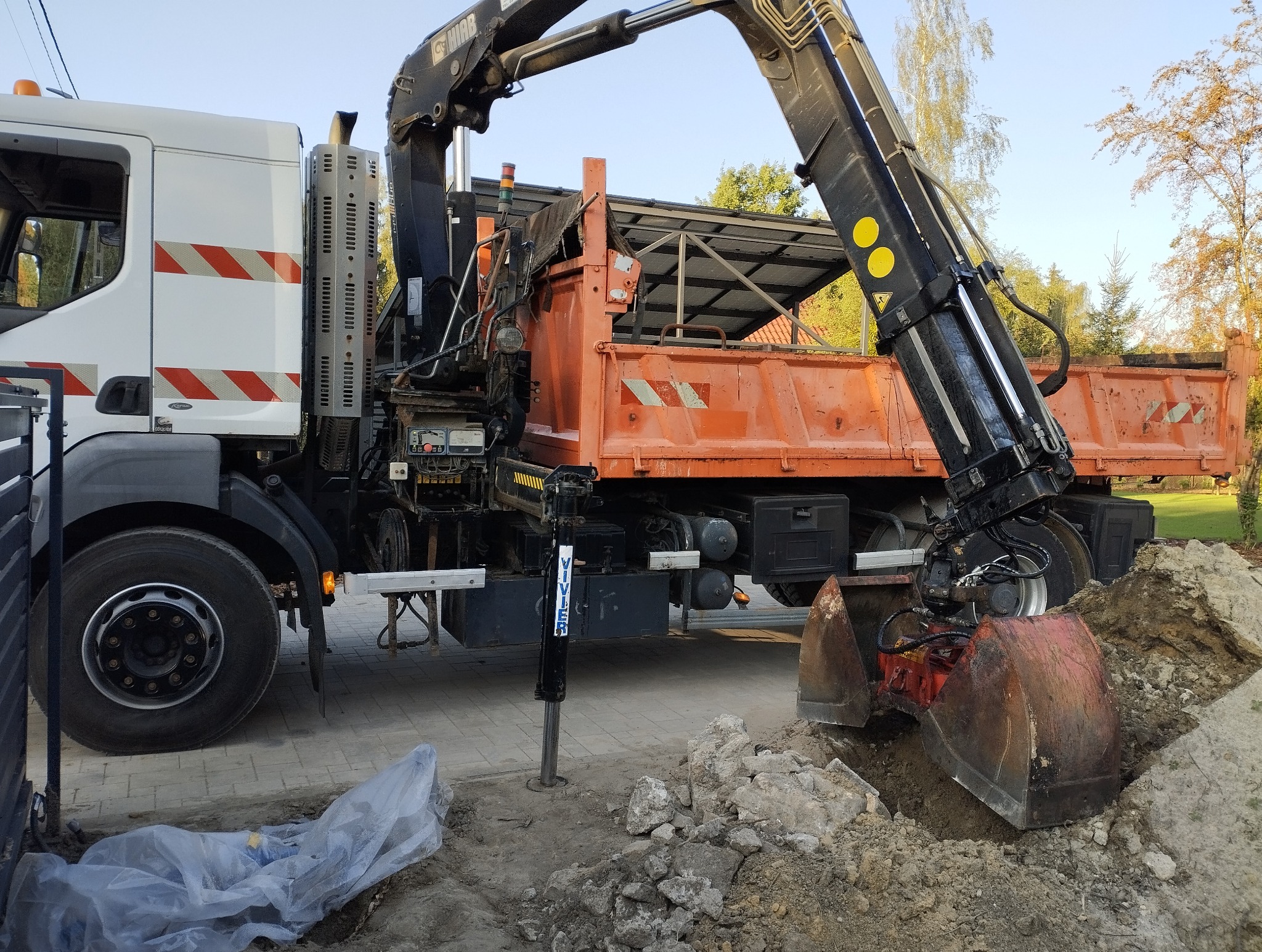 Pomarańczowa wywrotka z dźwigiem kopie ziemię na działce. Widoczne podpory stabilizujące pojazd. Ziemia w wykopie i na pryzmie. Częściowo widoczne ogrodzenie.