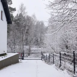 Zimowy krajobraz z metalową bramą wjazdową i siatką ogrodzeniową, pokryte śniegiem, prowadzące do nowoczesnego, białego domu z balkonem.