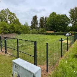 Nowoczesne ogrodzenie panelowe z siatki w Gdyni, otaczające duży trawnik z drzewami w tle, widok z poziomu skrzynki elektrycznej.