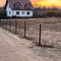 FIRMA HANDLOWO-USŁUGOWA THOMAS TOMASZ KOWALIK - Nowoczesne, czarne ogrodzenie panelowe z siatki na tle domu jednorodzinnego i pomarańczowego nieba o zachodzie słońca. Widok z perspektywy gruntu.