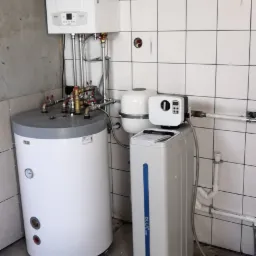 Kocioł gazowy wiszący nad zasobnikiem ciepłej wody i stacją uzdatniania wody w pomieszczeniu z częściowo wykończonymi ścianami i podłogą.