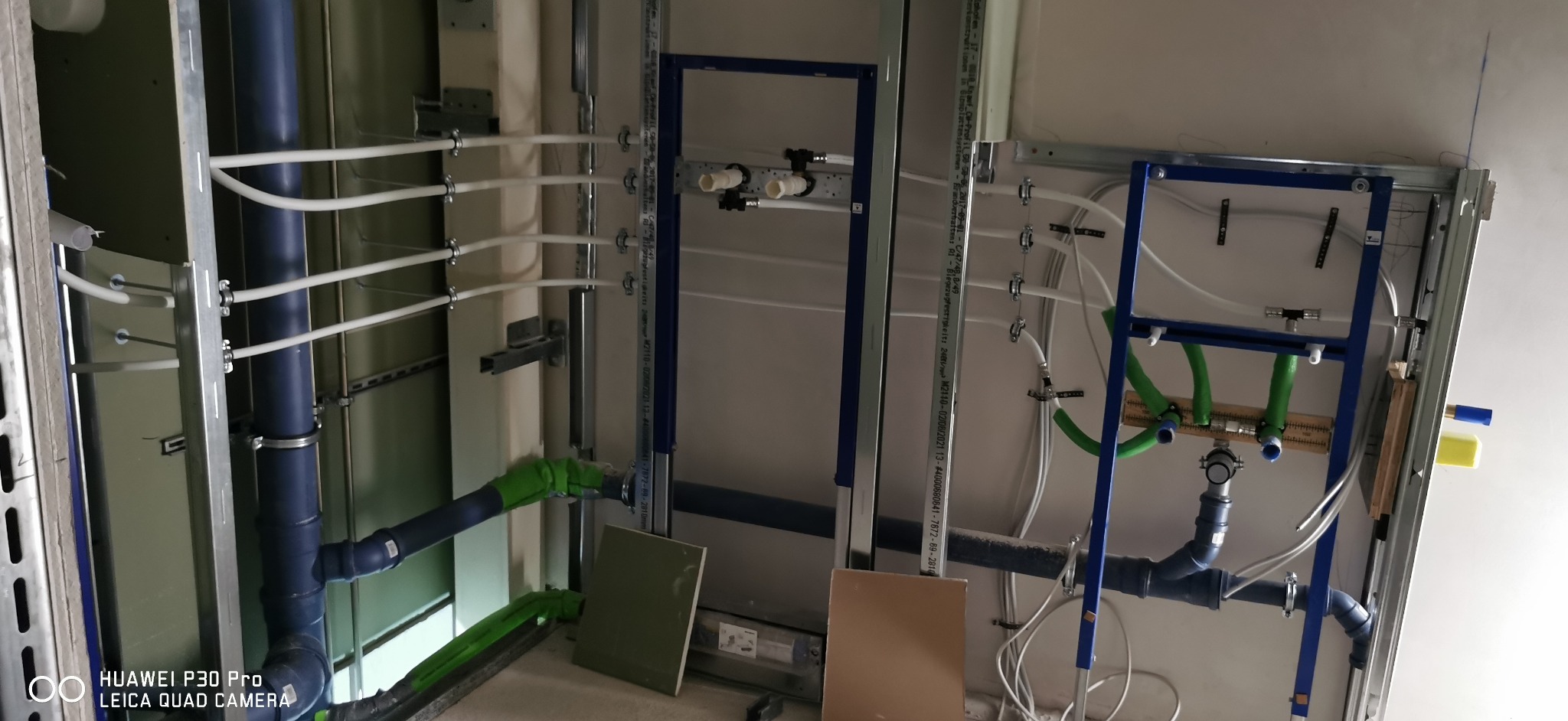 Wewnętrzna instalacja wodno-kanalizacyjna w trakcie montażu, widoczne rury PCV w kolorze niebieskim i białym, metalowe stelaże montażowe i elementy mocujące do ściany.