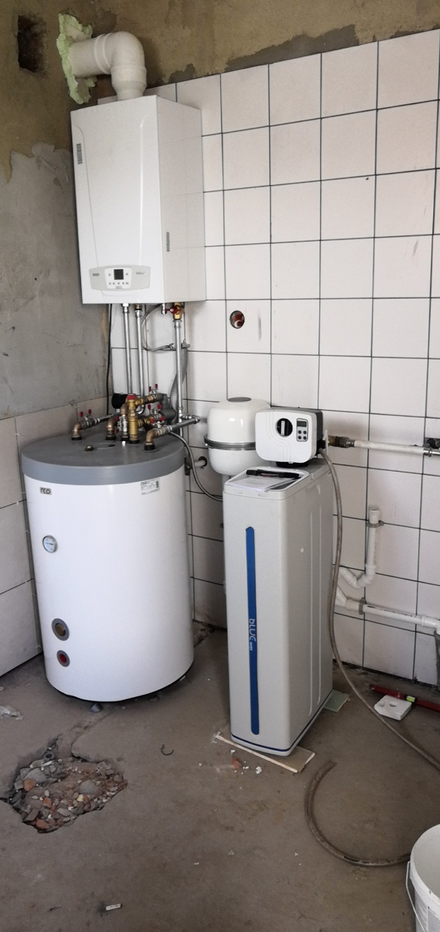 Kocioł gazowy wiszący nad zasobnikiem ciepłej wody i stacją uzdatniania wody w pomieszczeniu z częściowo wykończonymi ścianami i podłogą.