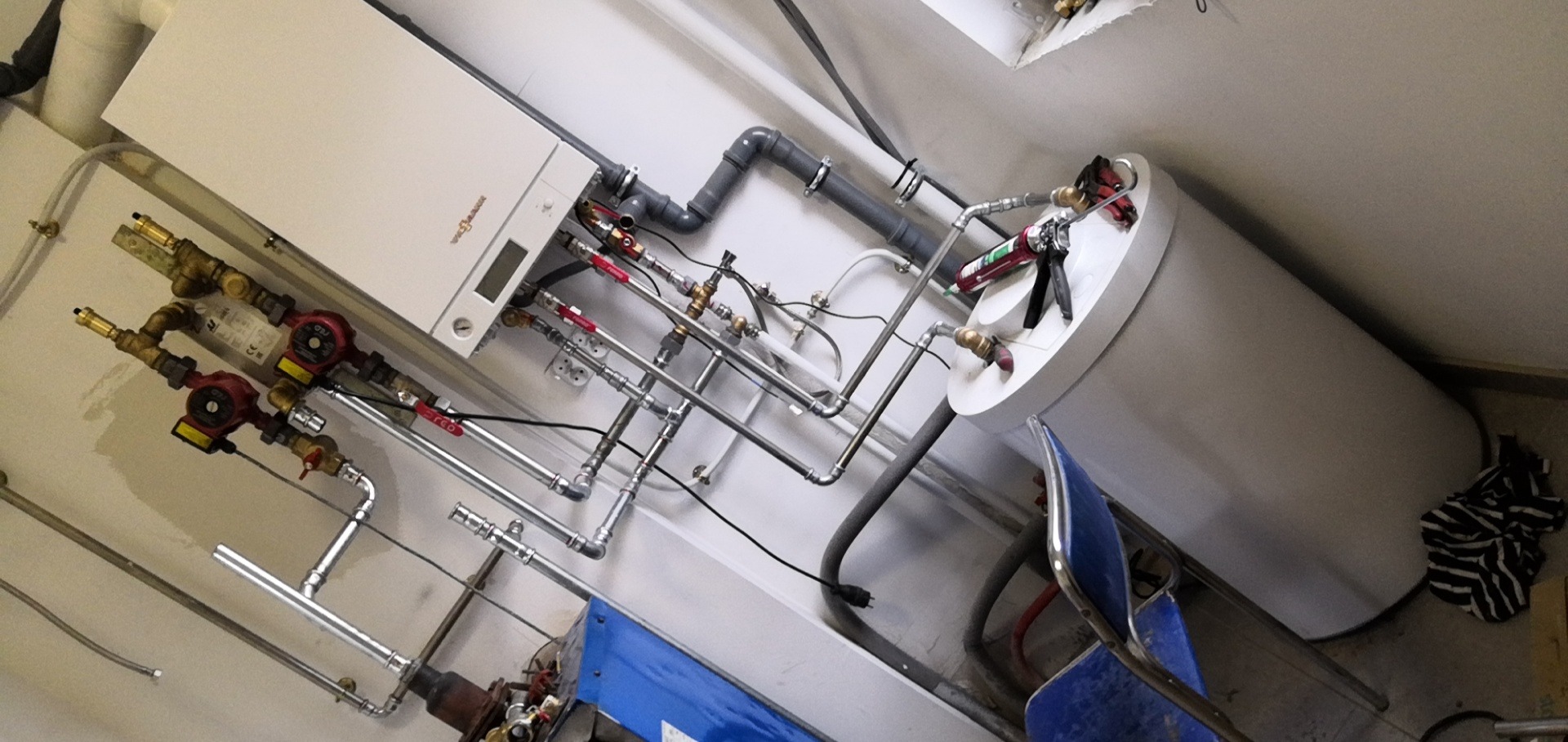 Biały piec gazowy Viessmann z podłączonymi rurami miedzianymi i pompami obiegowymi, obok biały zasobnik ciepłej wody, widoczny aplikator silikonu, ujęcie pod kątem.