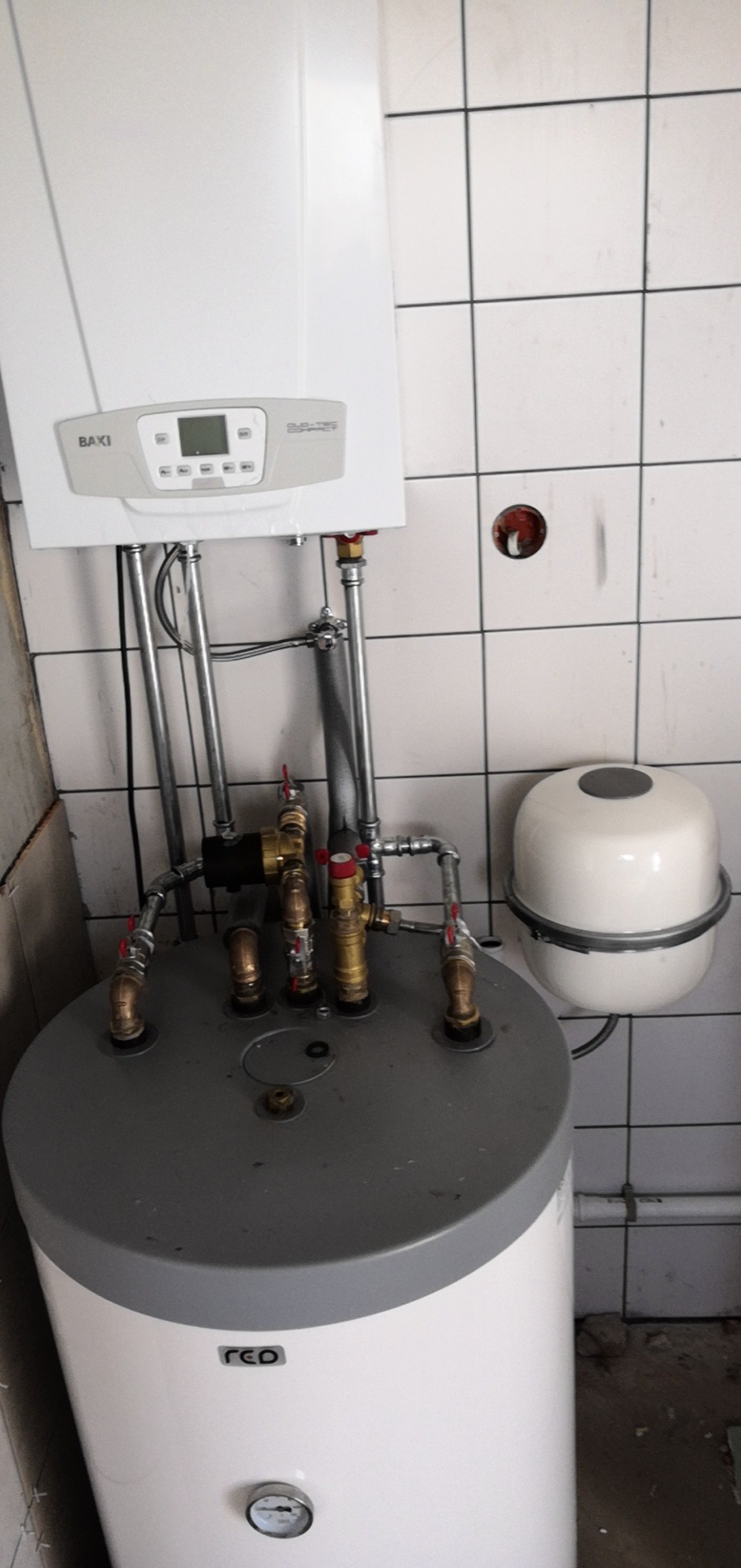 Biały piec gazowy marki Baxi zamontowany nad szarym zasobnikiem wody, z widocznymi rurami i zaworami, na tle białych płytek ceramicznych.