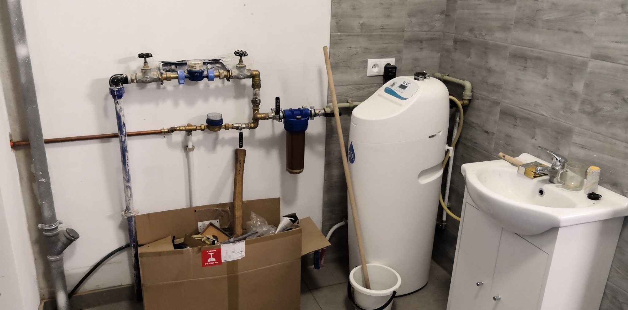 Biały zmiękczacz wody 20L zainstalowany w pomieszczeniu gospodarczym, widoczne rury, filtry, zawory, skrzynka z narzędziami i umywalka z akcesoriami malarskimi.