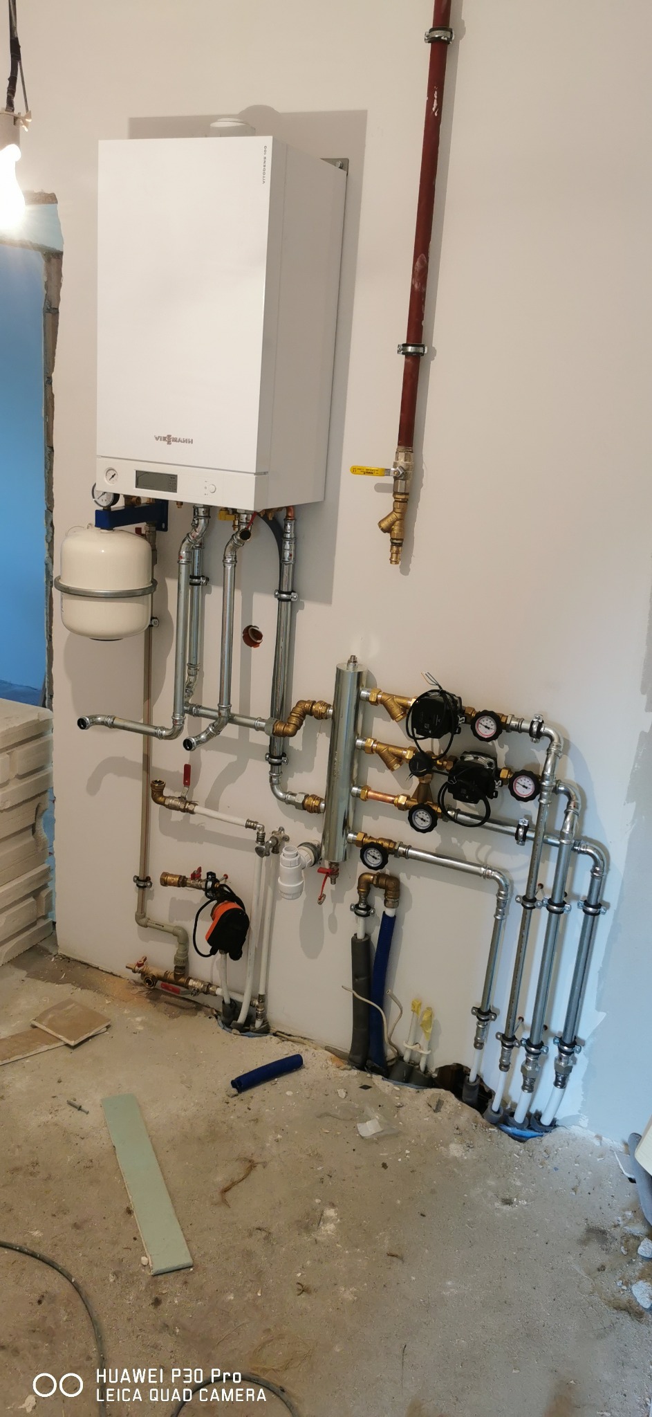 Biały piec gazowy Viessmann zamontowany na ścianie z licznymi rurami miedzianymi i stalowymi, manometry, zawory, instalacja w surowym stanie, widoczne ślady po kuciu w ścianie i na podłodze.