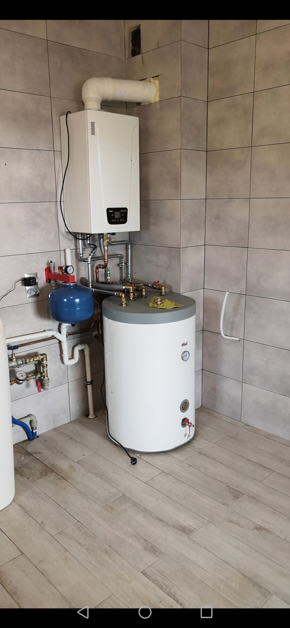 Nowoczesny piec gazowy wiszący marki Baxi z podłączonym zasobnikiem ciepłej wody Red, zainstalowany w pomieszczeniu wyłożonym szarymi płytkami i drewnopodobną podłogą.