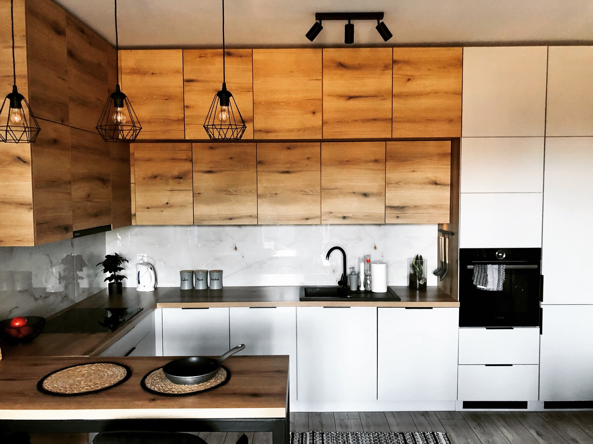 Nowoczesna kuchnia z drewnianymi szafkami górnymi, białymi szafkami dolnymi, czarnym zlewem i wiszącymi lampami w stylu industrialnym.