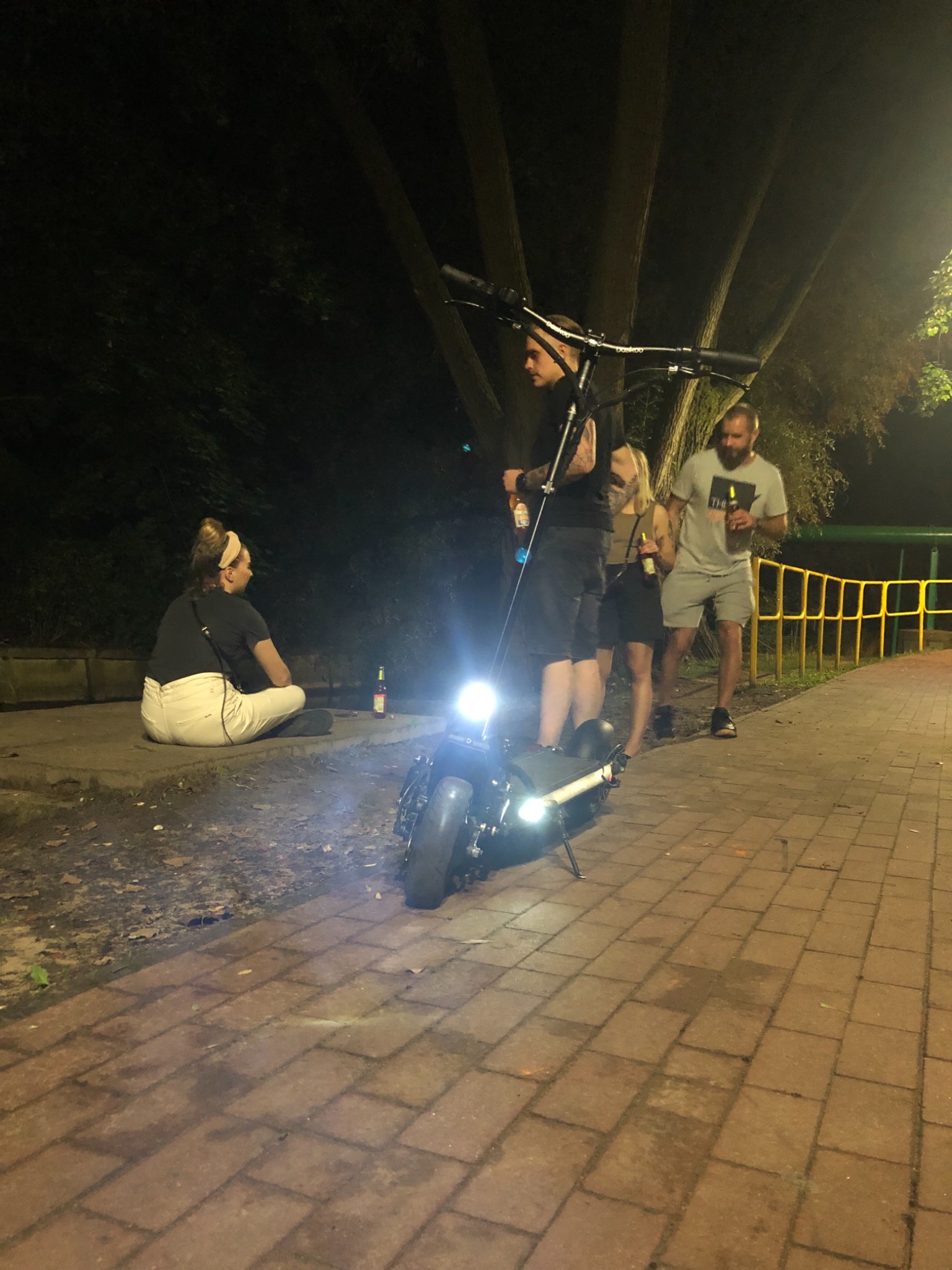 Nocne spotkanie towarzyskie na chodniku z elektryczną hulajnogą o mocnym oświetleniu, w tle drzewa i żółta barierka.