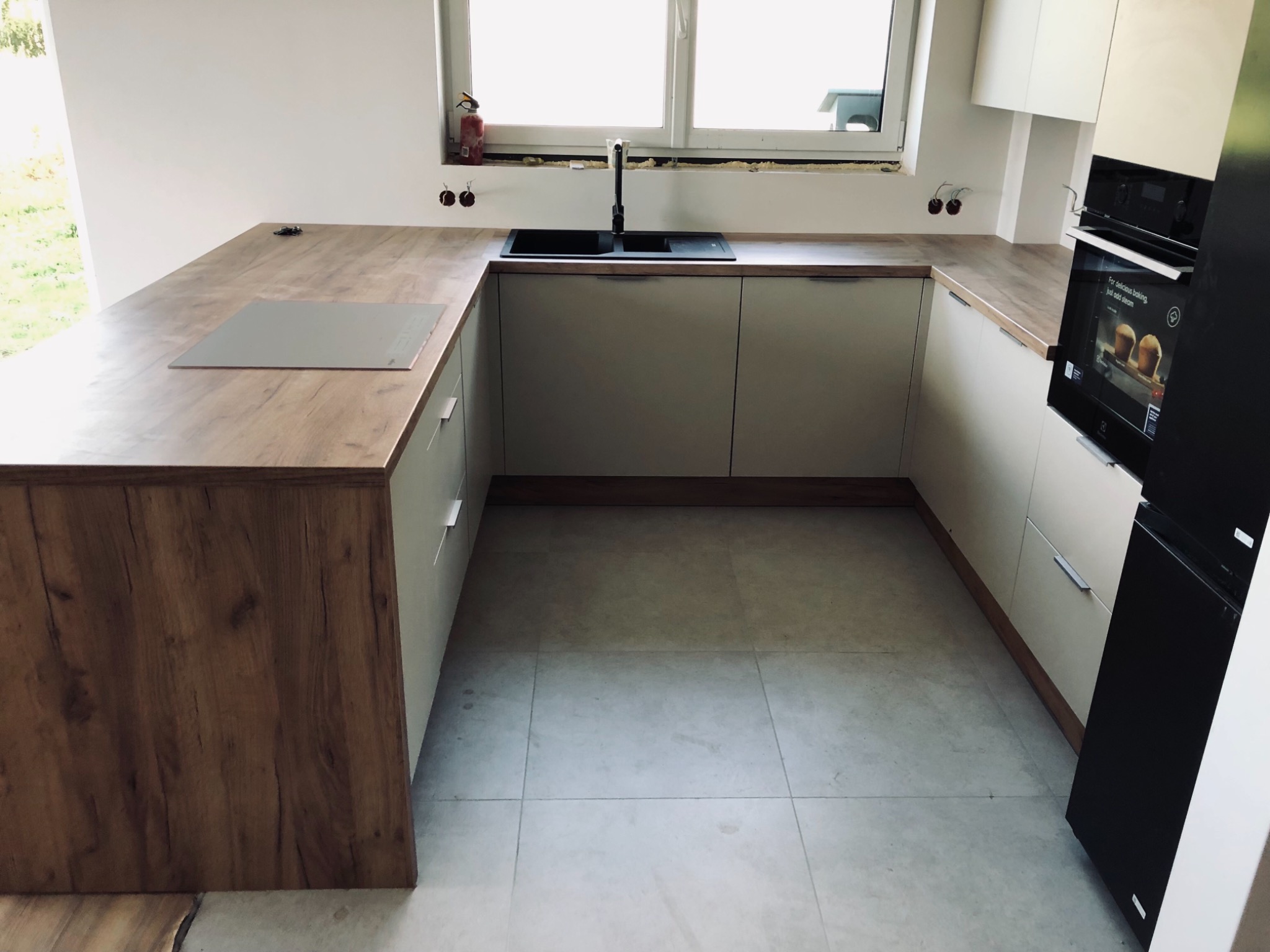 Nowoczesna kuchnia w kształcie litery U z drewnianym blatem, czarnym zlewem i płytą indukcyjną, z piekarnikiem z funkcją pary i czarną lodówką, widok na okno z widocznymi elementami instalacji.