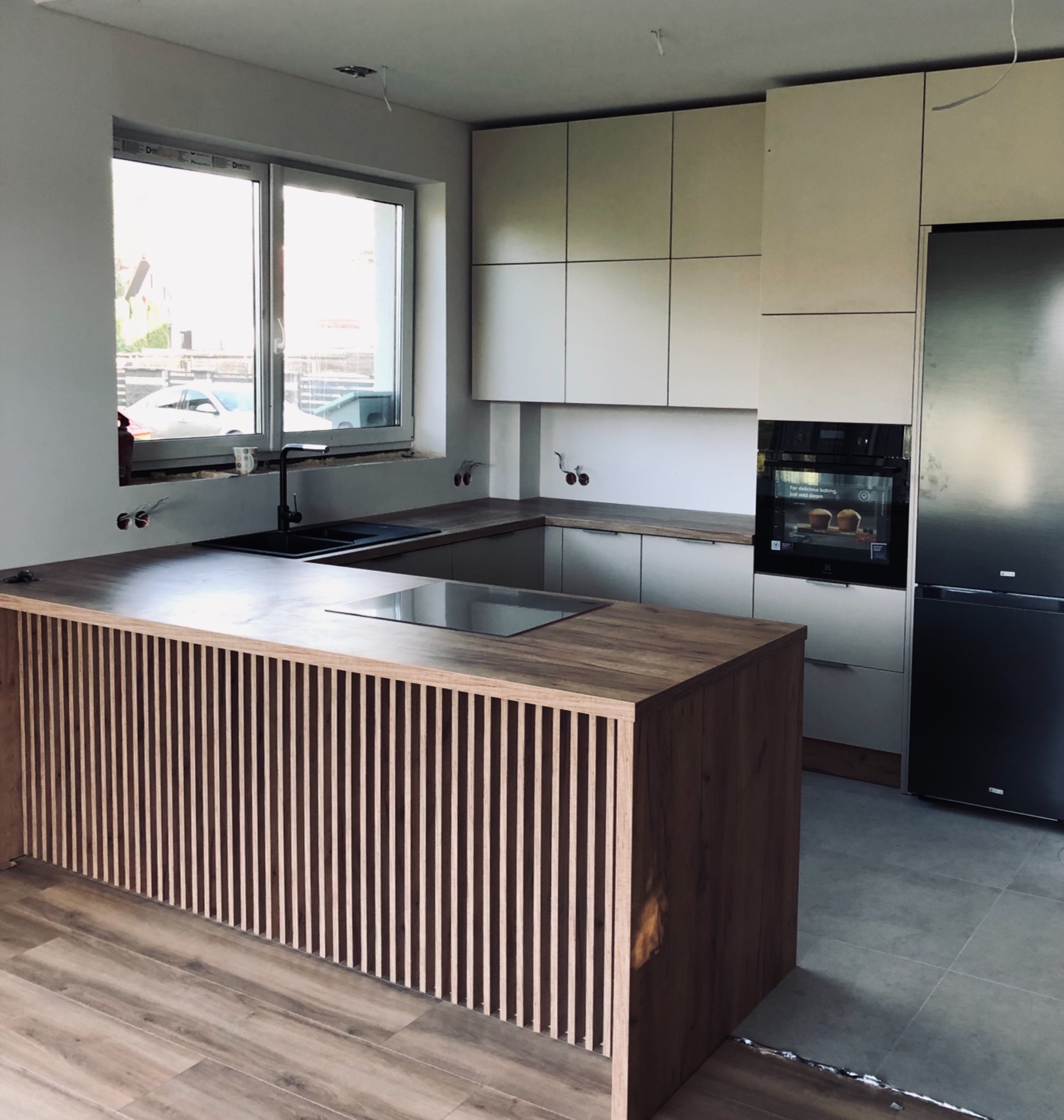 Wnętrze kuchni w trakcie wykańczania z wyspą z drewnianymi lamelami, widoczne niedokończone instalacje elektryczne przy oknie, nowoczesne białe szafki i czarny zlew.