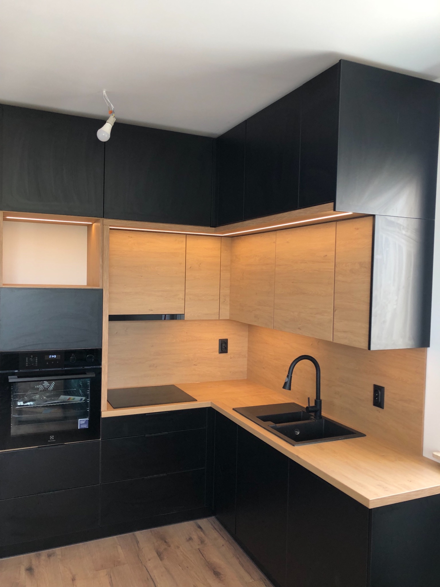 Nowoczesna kuchnia w odcieniach czerni i drewna, z oświetleniem LED podszafkowym, czarnym zlewem i baterią, oraz wiszącą żarówką bez klosza.