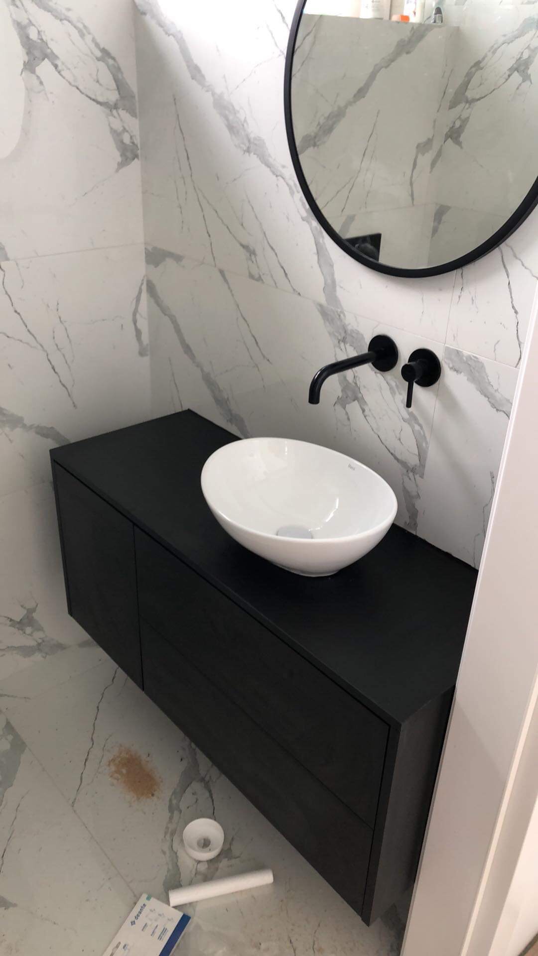 Nowoczesna łazienka z umywalką nablatową w kształcie misy, czarną szafką pod umywalką i okrągłym lustrem z czarną ramą na tle ściany wyłożonej płytkami imitującymi marmur.