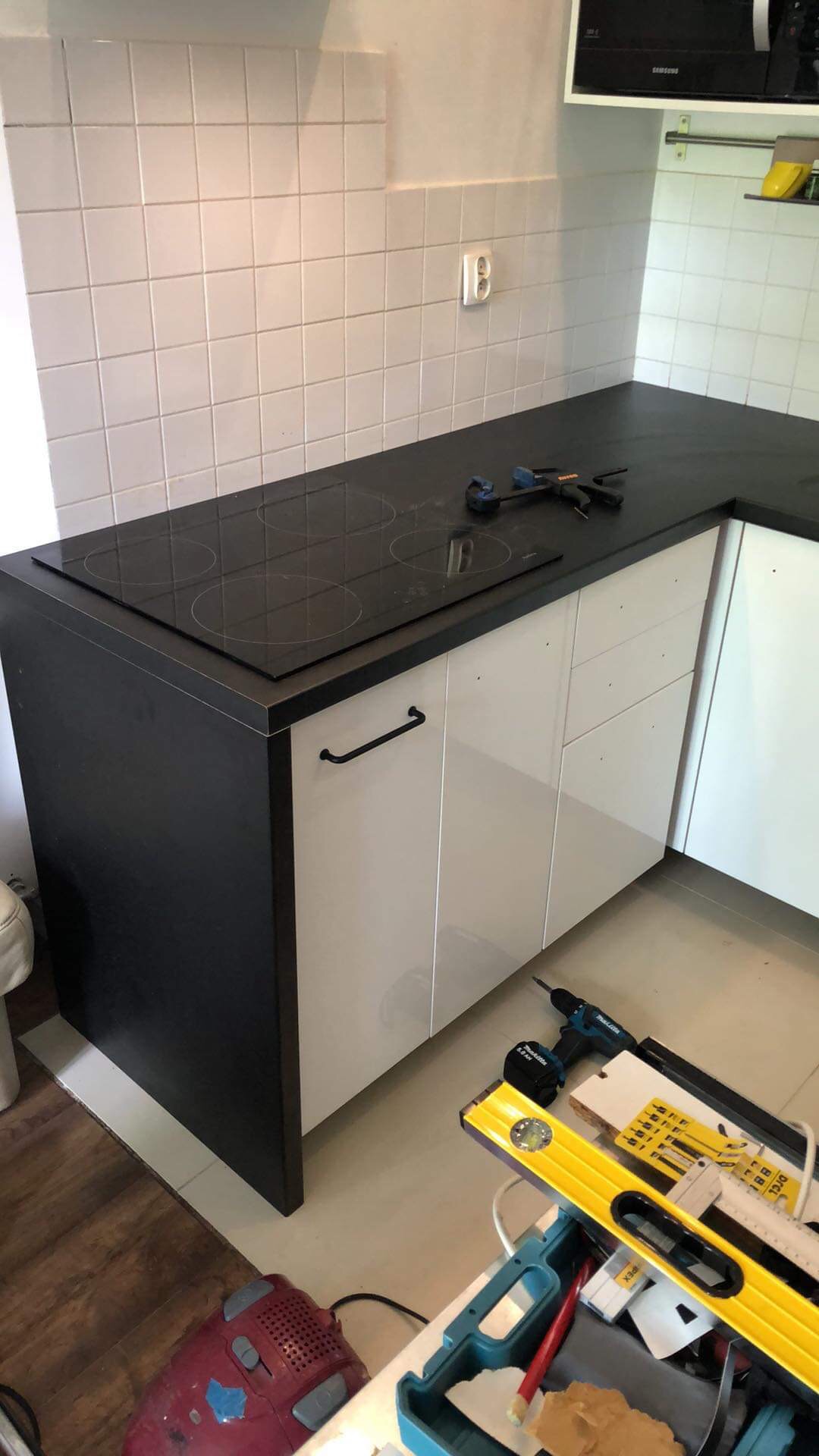 Fragment kuchni z czarnym blatem i płytą indukcyjną, białe fronty szafek, narzędzia na blacie i na podłodze w trakcie montażu.