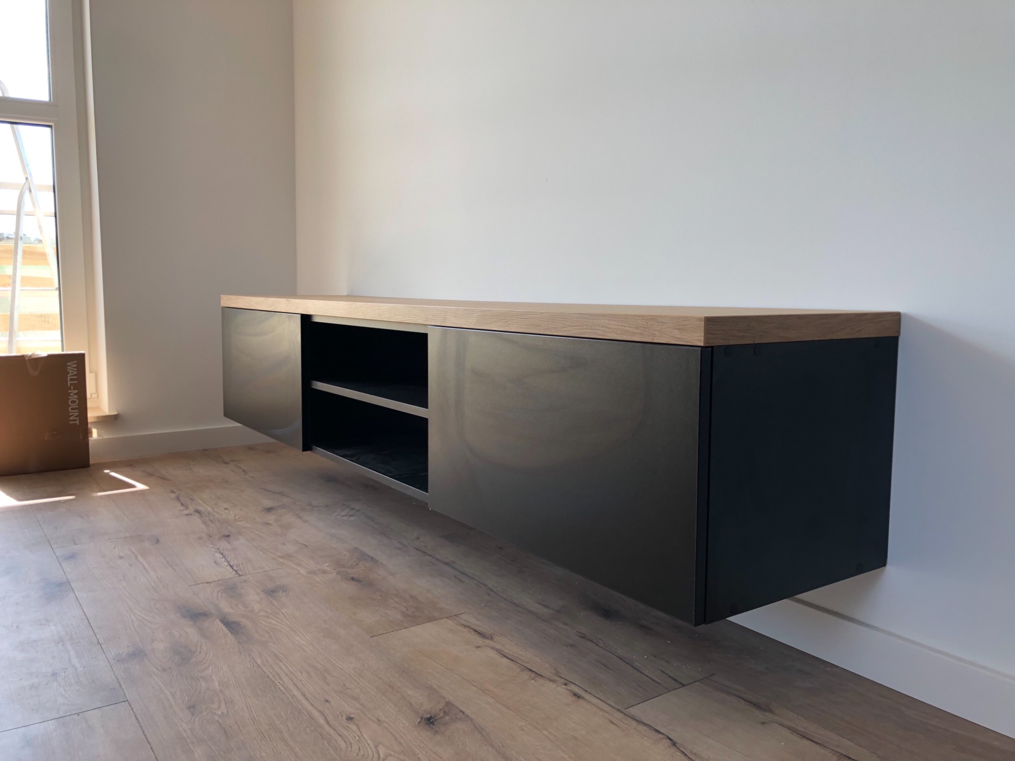 Czarna, wisząca szafka RTV z blatem z jasnego drewna zamontowana na białej ścianie, widoczna podłoga z paneli w odcieniach brązu i fragment okna z kartonem w rogu.