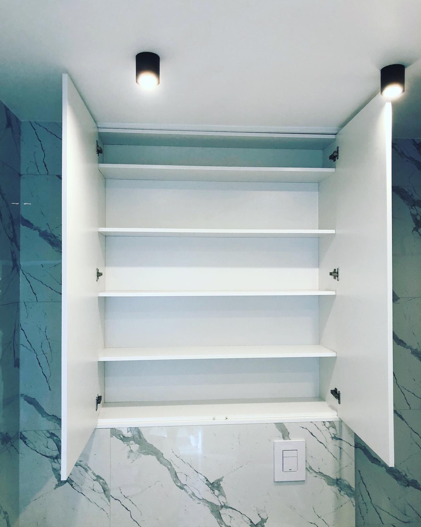 Biała szafka wisząca z otwartymi drzwiczkami i półkami, zamontowana na ścianie wyłożonej marmurowymi płytkami, oświetlona dwoma punktowymi lampami sufitowymi.