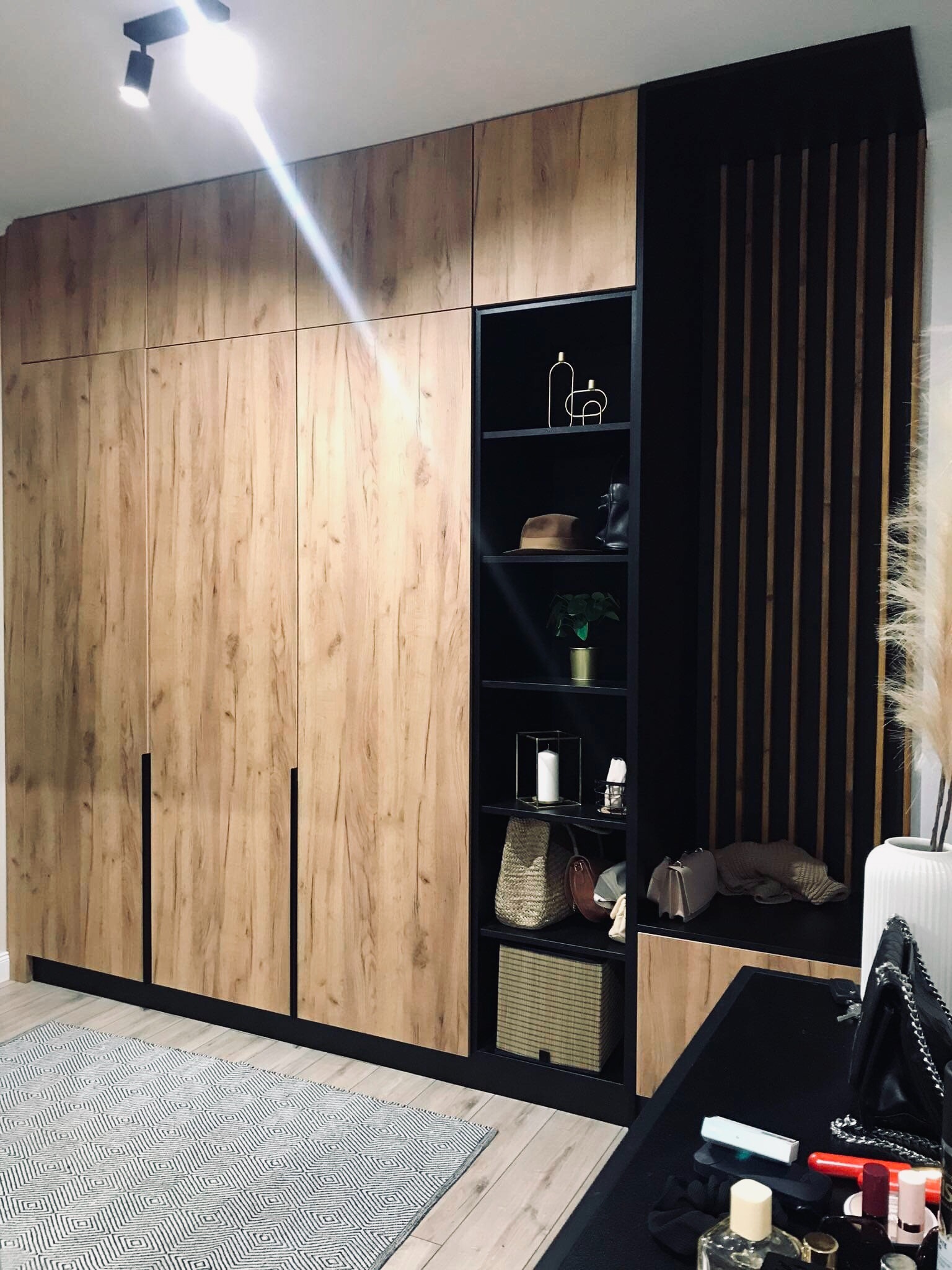 Duża szafa z drewnianymi frontami i czarnymi akcentami, z otwartymi półkami z dekoracjami, na podłodze wzorzysty dywan.